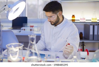 Scientist Preparing Research Paper Article For Modern Medical Journal. Portrait Of Serious Man In Lab Coat And Goggles Sitting At Table In Science Laboratory, Reading Scientific Text And Thinking