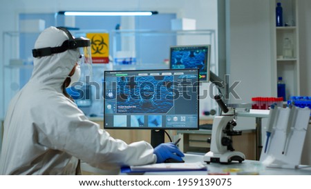 Scientist in protection suit typing on pc working in medical laboratory, writing on clipboard, developing inovative drugs against new virus. Doctor using high tech, chemistry tools for researching