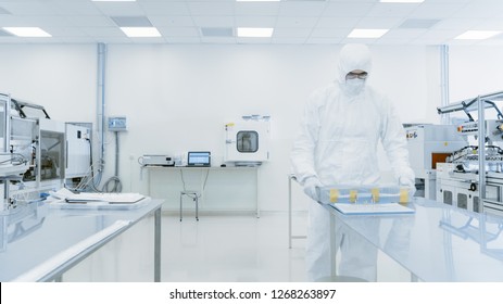 Scientist In Protective Suit Working With Case Of Finished Product Through Laboratory. Workers In The Facility With Modern Industrial Machinery. Product Manufacturing Process: Pharmaceutics, Biotech.