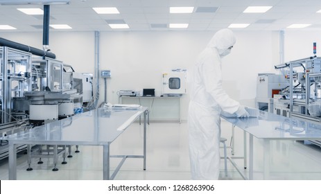 Scientist In Protective Suit Working With Case Of Finished Product Through Laboratory. Workers In The Facility With Modern Industrial Machinery. Product Manufacturing Process: Pharmaceutics, Biotech.