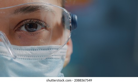 Scientist Showing One Eye On Camera In Laboratory, Wearing Safety Glasses And Face Mask. Woman Biologist Looking At Camera With Face Of Face, Working In Research Lab. Close Up