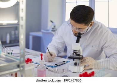 Scientist Taking Notes On Paper Notepad Holder. Serious Man In Lab Coat And Goggles Writing Scientific Research Data Analysis Report Sitting At Table With Microscope In Microbiology Science Laboratory