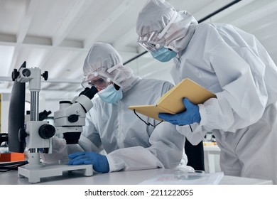 Scientist Team, Covid Research And Microscope With Team Writing Notes Or Results During Analysis Of A Sample In A Science And Medical Lab. Medical Researcher In Safety Suit In Biotechnology Laborator