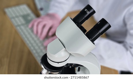 Scientist uses microscope to examine samples in a laboratory setting with computer keyboard nearby
