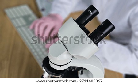 Scientist uses microscope to examine samples in a laboratory setting with computer keyboard nearby
