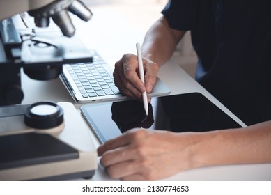 Scientist using digital tablet and working on laptop computer with microscope. Doctor or technician recording lab test on tablet pc in hospital laboratory, close up, medical research concept