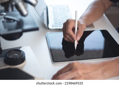 Scientist using digital tablet and working on laptop computer with microscope. Doctor or technician recording lab test on tablet pc in hospital laboratory, close up