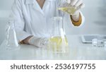 A scientist, wearing a lab coat and white protective gloves, is pouring a yellow oily liquid from one beaker to another in laboratory, close up. Medicine and science concept