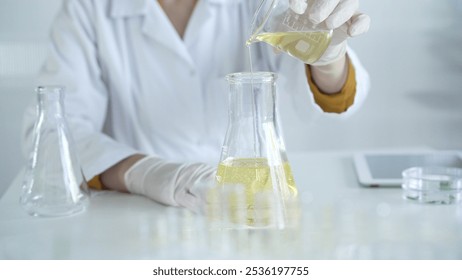 A scientist, wearing a lab coat and white protective gloves, is pouring a yellow oily liquid from one beaker to another in laboratory, close up. Medicine and science concept