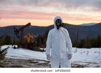 Scientist Wearing White Coverall, Gloves And A Gas Mask Working In Oil Field In Mountainous Region, On Background Oil Rig Against Purple Sunset. Concept Of Ecology, Environmental Pollution.