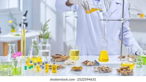 A scientist in a white lab coat pours yellow liquid from a flask into a funnel in a laboratory setting. There are various dried herbs, flasks, and test tubes in the background. - Powered by Shutterstock