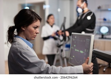 Scientist woman analyzing heart rate pulse on monitor while in background athlete man with electrodes working at body endurance running on fitness trainer in lab. Specialist doctor monitoring EGK data - Powered by Shutterstock