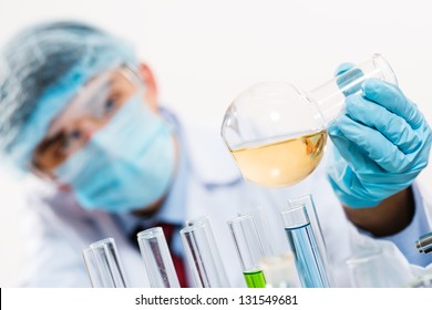 Scientist Working In The Lab, In Protective Mask And Cap, Examines A Test Tube With Liquid