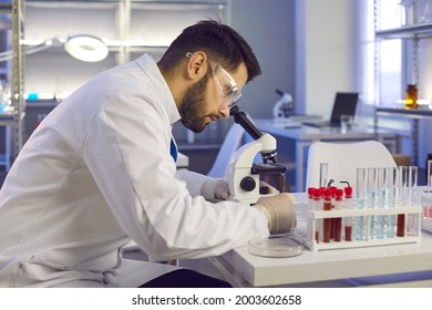 Scientist Working In Modern Pharma, Biotech Or Microbiology Science Laboratory. Side View Profile Portrait Of Serious Man In Lab Coat Sitting At Table With Glassware Looking At Sample Under Microscope