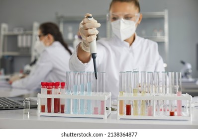 Scientist Working In Modern Pharma Or Biotech Laboratory. Serious Woman In PPE Mask And Goggles Doing Research And Using Pipette To Transfer Liquid In Glass Tubes. Soft Focus Close Up, Science Concept