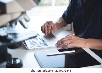 Scientist Working On Laptop Computer And Using Digital Tablet With Microscope. Doctor Or Technician Recording Lab Test On Laptop Computer In Hospital Laboratory, Close Up