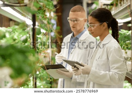Scientists studying and recording data on plant growth in a well-lit, controlled environment laboratory using digital tablets and wearing lab coats and safety glasses