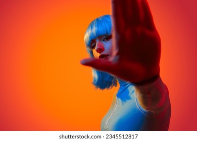 Sci-fi young female in trendy outfit and blue wig looking at camera and showing futuristic gesture against neon orange background - Shutterstock ID 2345512817