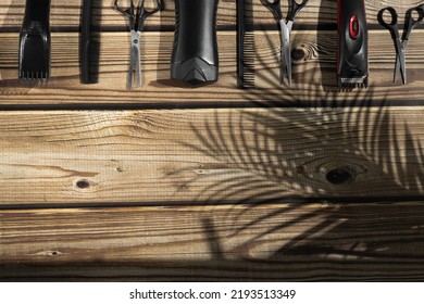 Scissors, Combs, Shampoo And Men's Hair Clippers Lie In A Row From Above On A Brown Wooden Background With Copy Space Below And A Shadow From A Palm Branch On The Right, Flat Lay Close-up.