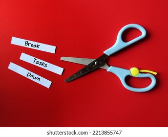 Scissors on red background, cut paper into pieces with handwritten text - Break Tasks Down - means to break down tasks into smaller parts which easier to work on, focus on doing one work at a time