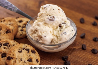 Scoop of chocolate chip cookie dough ice cream in small glass dish next to chocolate chip cookies and chocolate chips on wooden table