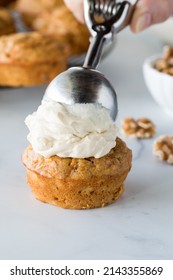 A Scoop Of Cream Cheese Frosting Being Placed On Top Of A Carrot Cake Muffin.