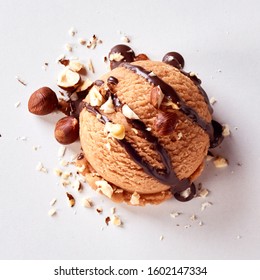 Scoop Of Creamy Hazelnut Ice Cream Topped With Fresh Chopped Nuts And Drizzled With Chocolate Sauce Over White In An Overhead View