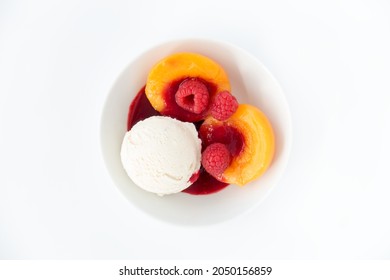 Scoop Of Homemade Vanilla Ice Cream In A White Bowl With Peach Halves, Fresh Raspberries And Raspberry Sauce Isolated On White.