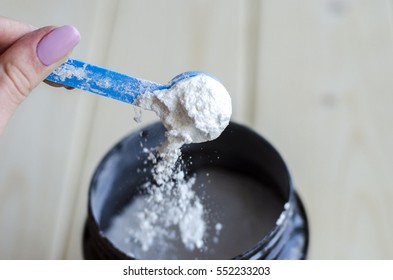 Scoop of white whey isolate protein in a black plastic container on white wooden background. Picture powder (sports nutrition, proteins) spilled from the measuring spoon, photo on the move