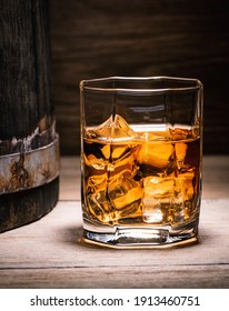 Scotch Whiskey, Rum, Or Bourbon. Glass Of Whiskey With Ice Cubes Sits On A Wooden Table Next To A Wooden Whiskey Barrel In The Basement Of A Wine Cellar.