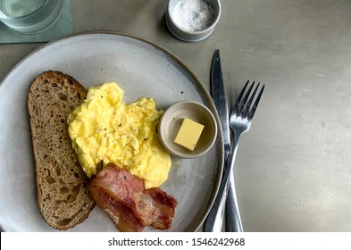 Scrambled Eggs,bacon, Toast, And Butter On Gray Plate,glass Water, Salt Ramekin And Concrete Counter Top With Natural Lighting Isolated With Room For Copy.