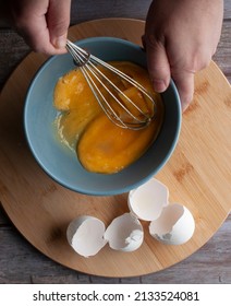 Scrambled Raw Eggs In A Blue Bowl Over A Round Wooden Chopping Board And Egg Shells On The Side, Overhead Lay Flat Or Top View, Vertical Image