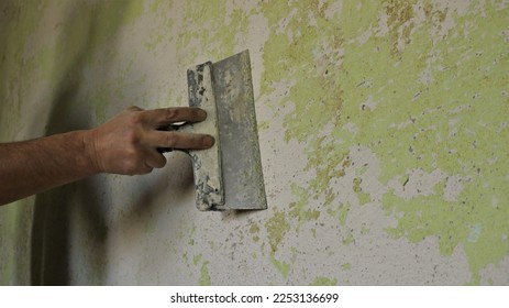 scraping off unevenness and residues of paint and plaster with a spatula, cleaning and preparing the surface of a room wall before sticking wallpaper, a hand scrapes off a layer on a dilapidated wall
