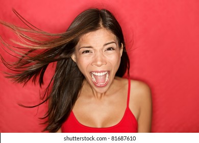 Scream. Woman screaming wild and crazy at full energy looking at camera on red background. Beautiful mixed race Asian Caucasian brunette female model with wind in the hair.