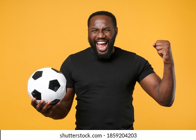 Screaming african american man 20s football fan in black t-shirt isolated on yellow background. Sport family leisure lifestyle concept. Cheer up support favorite team with soccer ball, clenching fist