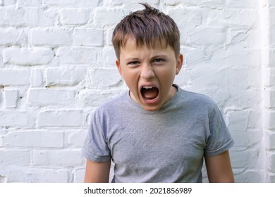 A Screaming Boy Teenager With His Mouth Wide Open And Squinted Eyes Against The Background Of A White Brick Stubble