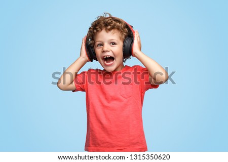 Screaming excited curly kid in headphones listening to music on blue background 