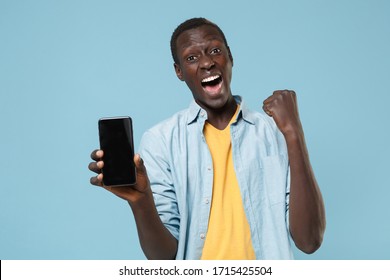 Screaming young african american man guy in casual shirt, yellow t-shirt posing isolated on blue background. People lifestyle concept. Hold mobile phone with blank empty screen doing winner gesture