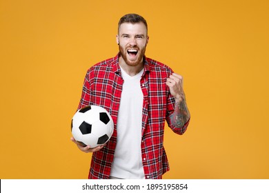 Screaming young man football fan in basic shirt cheer up support favorite team with soccer ball clenching fist doing winner gesture isolated on yellow background studio. People sport leisure concept