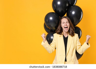 Screaming young woman in suit jacket celebrating birthday holiday party with bunch of air ballons clenching fists doing winner gesture isolated on yellow background studio portrait. Black friday sale Foto Stock
