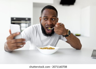 Screen Addiction. Portrait Of Black Guy Having Breakfast In Kitchen At Home, Eating Healthy Cereal, Using And Looking At Mobile Phone, Watching Movie Or Browsing Social Media Sitting At Dining Table