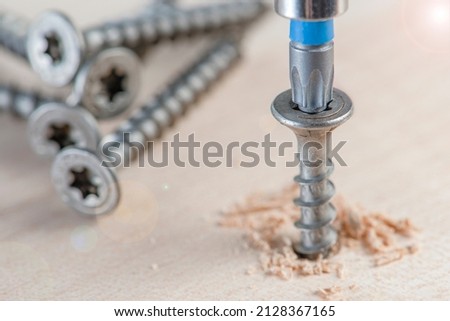 Screw the screw into the board. Furniture production, a screwdriver twists a self-tapping screw into a board. Several silver screws lie on the desktop