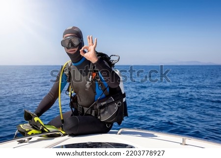 A scuba diver in full gear sits on a boat and signals the OK sign