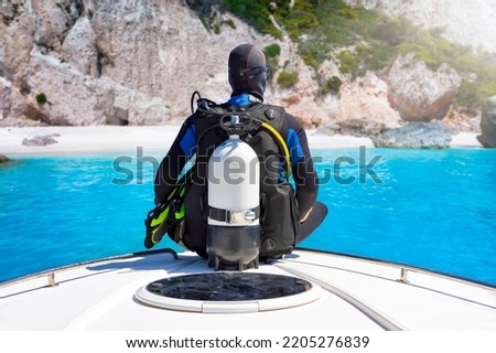 A scuba diver with his equipment sits on a bow of a boat in front of a beautiful beach with turquoise sea