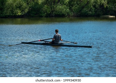 Scullers Sculling To Propel Their Long Narrow Boats Through The Water On A Lake Surrounded By Thick Vegetation And Green Trees. Some Crews Are Using Two Oars And Other Individuals Are Using One Oar