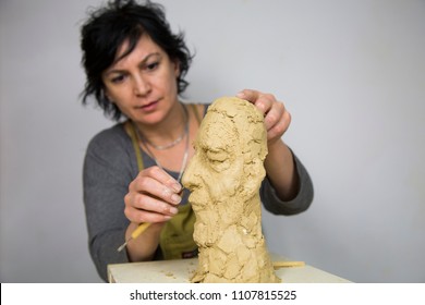 Sculptor artist creating a bust sculpture with clay. She is happy of her work, she is concentrated, she is sculpting a woman. the statue is at foreground. Sculptor at work.