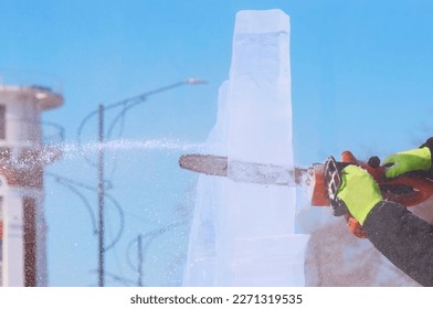 A sculptor carves a figure out of an ice block with an electric chainsaw against the backdrop of a lighthouse. Ice dust and ice shards in the air blur the background. Spring Festival on a sunny day.