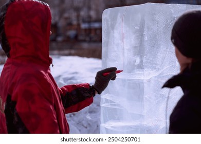 A sculptor draws a red line on an ice block. Preparing to create a sculpture at the city winter festival of artistic ice figures. A young guy in a red jacket with a hood. - Powered by Shutterstock