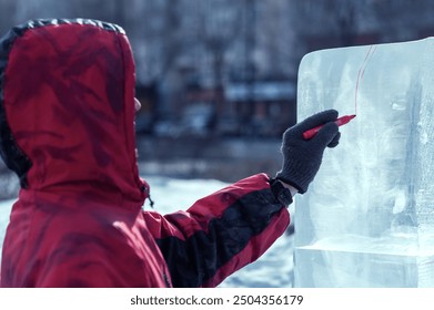 A sculptor draws a red line on an ice block. Preparing to create a sculpture at the city winter festival of artistic ice figures. A young guy in a red jacket with a hood. - Powered by Shutterstock