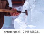 A sculptor processes a large ice cube with a sharp hand tool. Ice dust and ice fragments in the air. Ice sculpture festival in the city. Concept of hobby and interaction between man and nature.